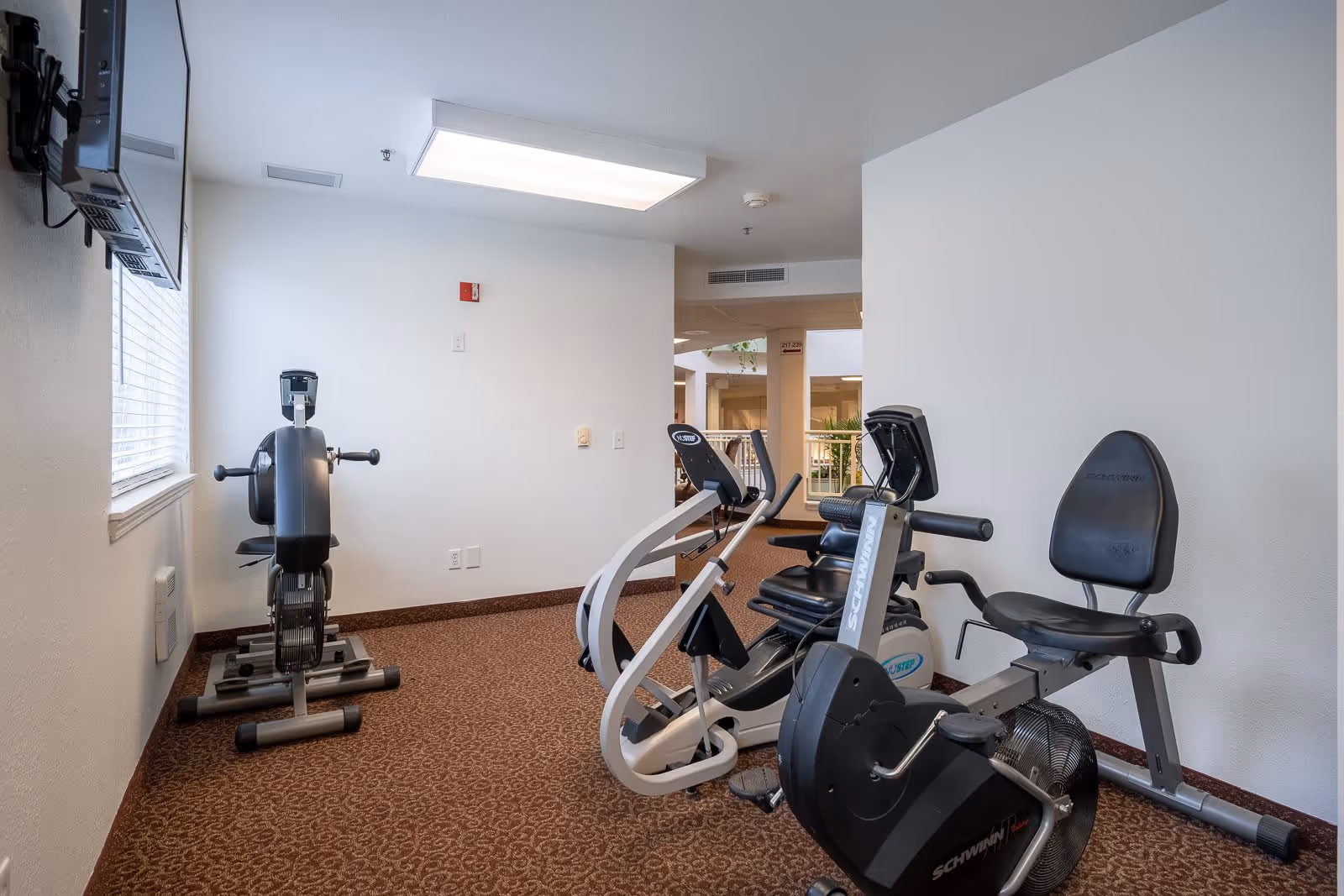 A small exercise room with stationary fitness equipment including a recumbent bike and an arm exercise machine. The room has white walls, a window with blinds, and a ceiling light panel. The floor is carpeted with a patterned brown carpet.