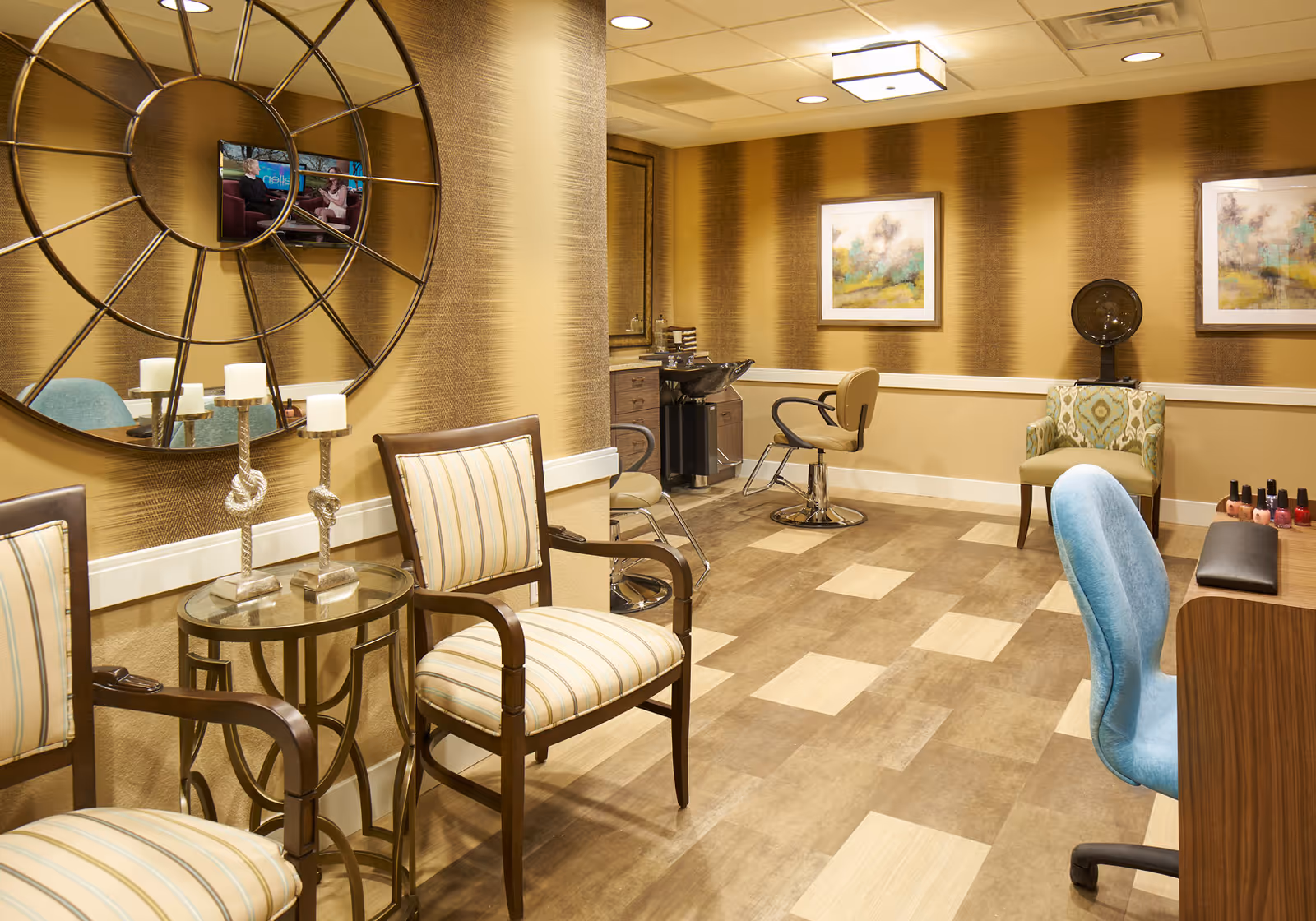 A well-lit salon room with striped beige and brown walls, featuring two striped armchairs around a small glass table with three candle holders, a large decorative round mirror, a hair washing station with a salon chair, a hair dryer chair, a blue office chair at a manicure table with nail polish bottles, and two framed abstract paintings on the wall.
