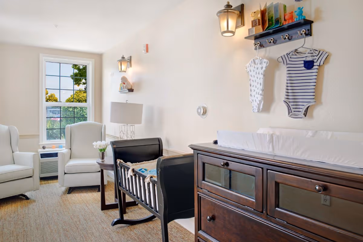 Bright interior sitting room with two armchairs, a wooden cradle, and a changing table beneath a wall shelf holding baby clothes.