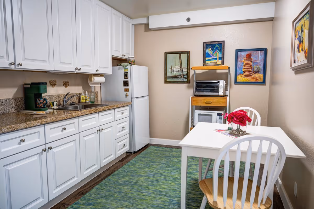 A small kitchen with white cabinets and granite countertops. There is a white refrigerator, a green coffee maker, a stainless steel sink, and a paper towel holder. On the right side, there is a white table with two white chairs, a vase with red flowers, and three colorful framed paintings on the beige walls. A green rug covers the floor.
