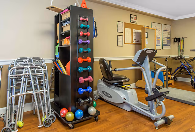 A senior facility fitness/therapy room with a recumbent exercise bike, a rack of colorful dumbbells, and stacked walkers.