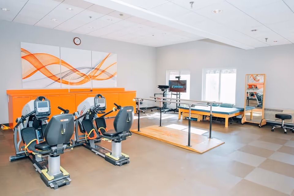 A rehabilitation and physical therapy room with exercise equipment including two recumbent bikes, parallel bars for walking practice, a therapy table, a mirror, and a stool. The room has bright lighting, a clock on the wall, and modern abstract artwork with orange and gray wave patterns above an orange cabinet.