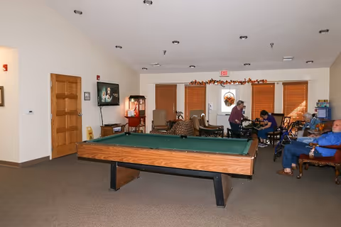 A common room in a senior living facility featuring a pool table in the center, several chairs and armchairs around the room, a television mounted on the wall, and a few elderly people sitting and interacting near the back. The room has beige walls, carpeted floor, and windows with wooden blinds.