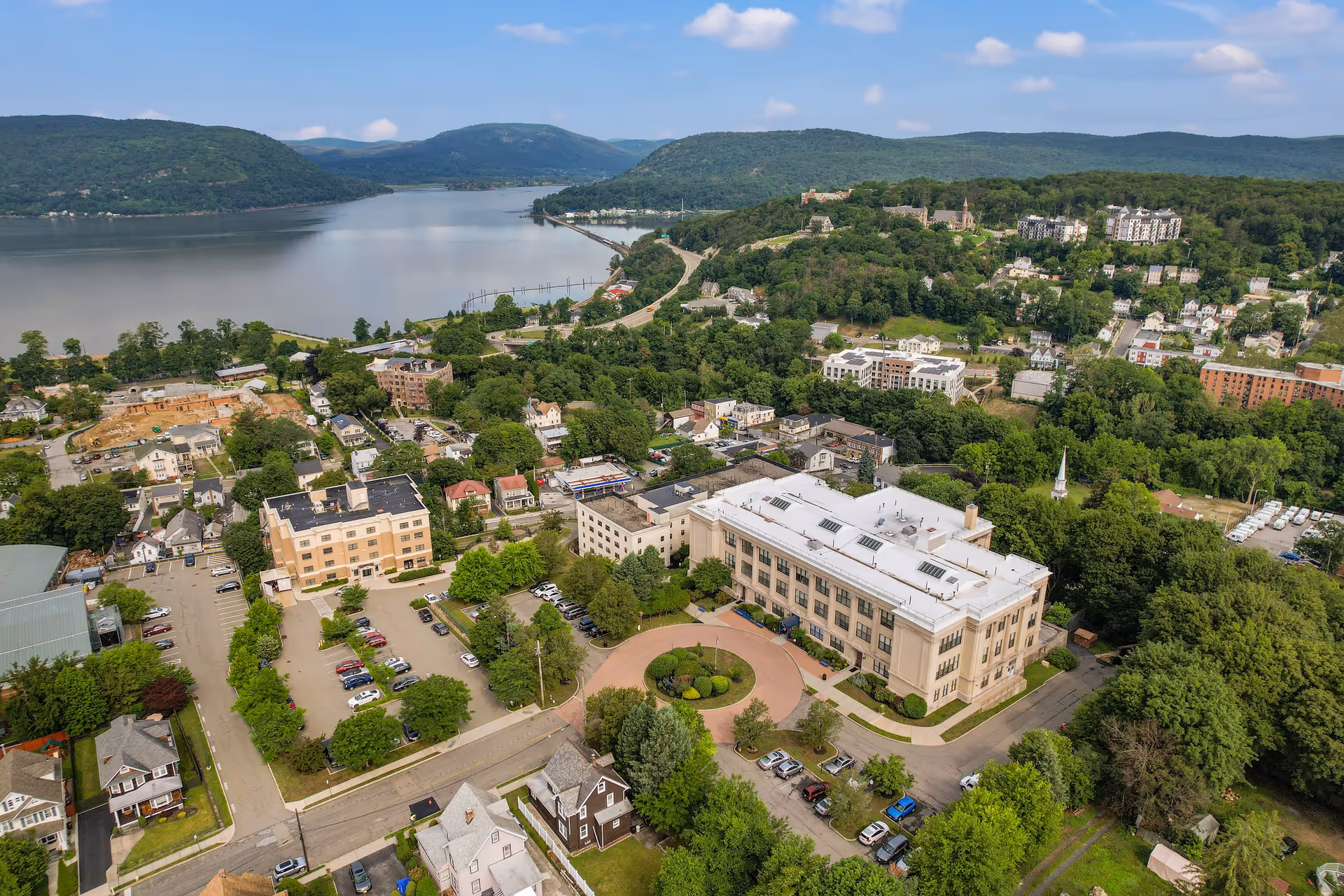 Aerial view of a senior living community surrounded by trees and residential houses, with a large river and hills in the background under a partly cloudy sky.