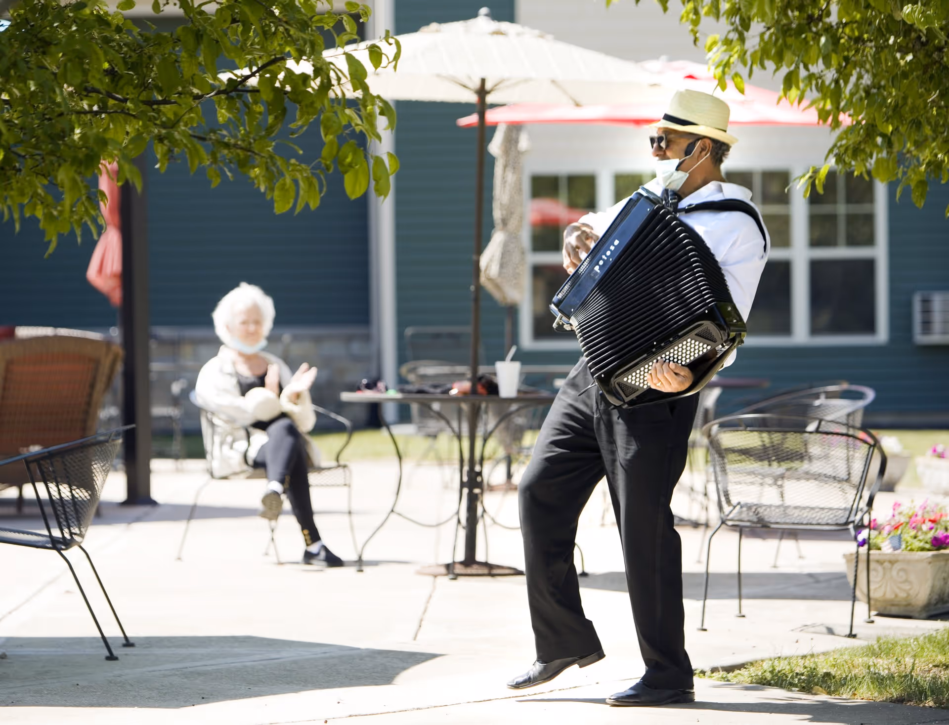 A man wearing a hat and sunglasses plays an accordion outdoors on a sunny day. In the background, an elderly woman sits on a chair clapping her hands. There are patio tables, chairs, umbrellas, and a building with windows behind them.