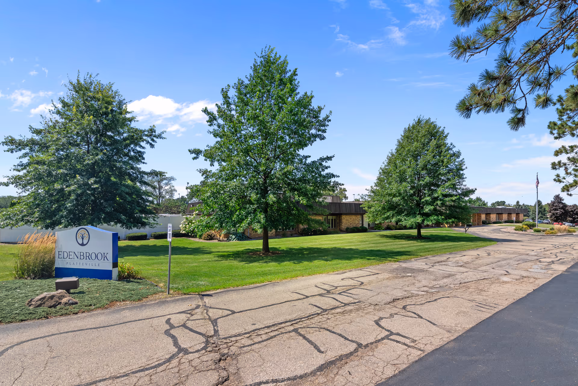Exterior view of Edenbrook Platteville facility with a paved driveway, green lawn, several trees, and a clear blue sky. A sign with the facility name is visible on the left side near the driveway entrance.