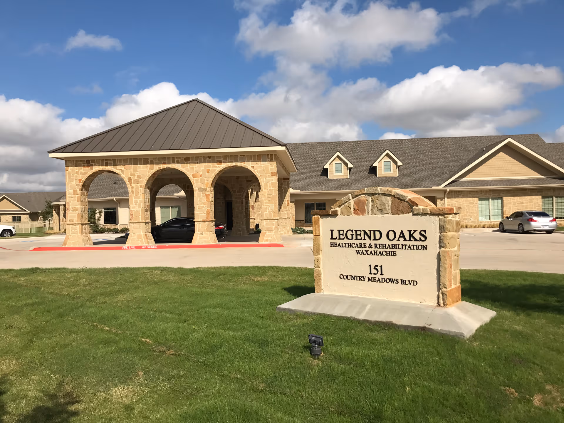 Exterior view of Legend Oaks Healthcare and Rehabilitation facility in Waxahachie, showing a stone entrance structure with arches, a building with a pitched roof in the background, a sign with the facility name and address, and a green lawn under a partly cloudy sky.