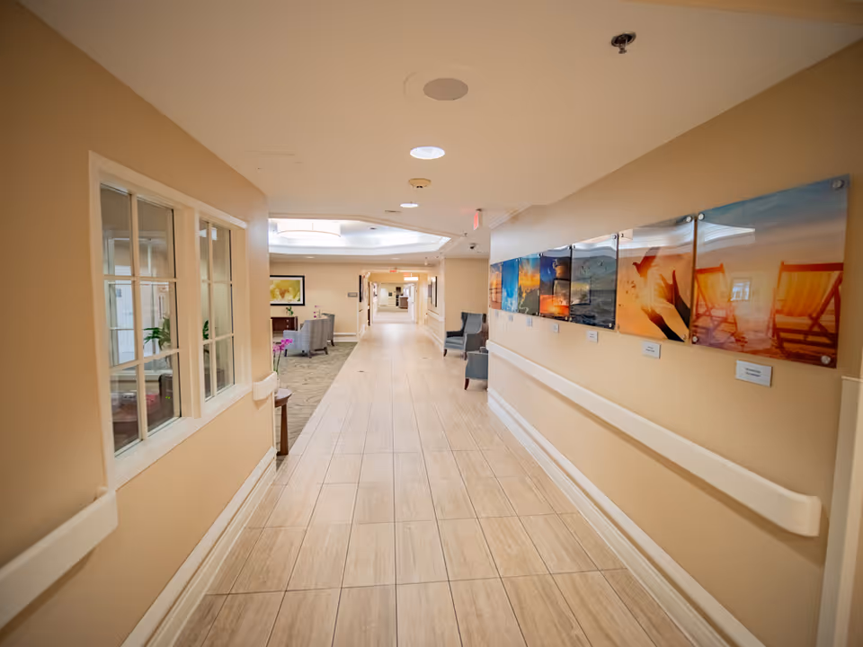 A well-lit hallway in Solaris Healthcare East Orlando with beige walls and light wood flooring. The right wall features a series of colorful framed pictures, and there are handrails along both sides of the hallway. At the far end, there are chairs and a seating area with a table and plants visible through a window on the left side.
