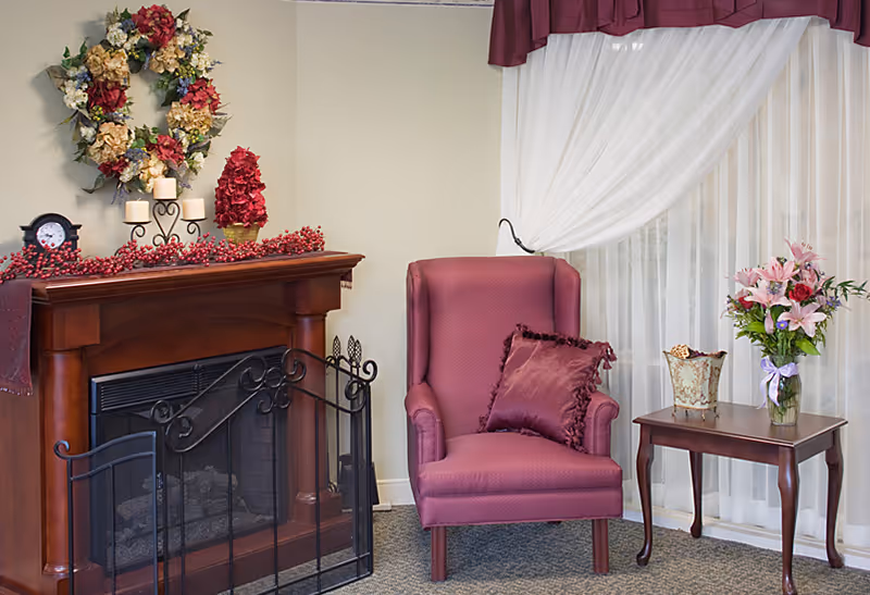 Cozy corner of a room featuring a wooden fireplace mantel decorated with candles, a floral wreath, and red berry garland. Next to the fireplace is a maroon upholstered armchair with a matching pillow, and a wooden side table holding a vase of pink lilies and a decorative container. Sheer white curtains and a maroon valance cover the window behind the chair and table.