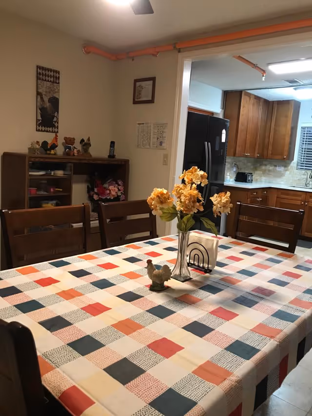 A dining area with a rectangular table covered by a colorful checkered tablecloth in shades of red, orange, blue, and cream. On the table is a vase with yellow flowers, a small ceramic rooster, and a napkin holder. Surrounding the table are wooden chairs. In the background, there is a kitchen with wooden cabinets, a black refrigerator, a toaster, and a sink. To the left, there is a wooden shelf with decorative items and flowers, and a wall with framed pictures and a ceiling fan above.