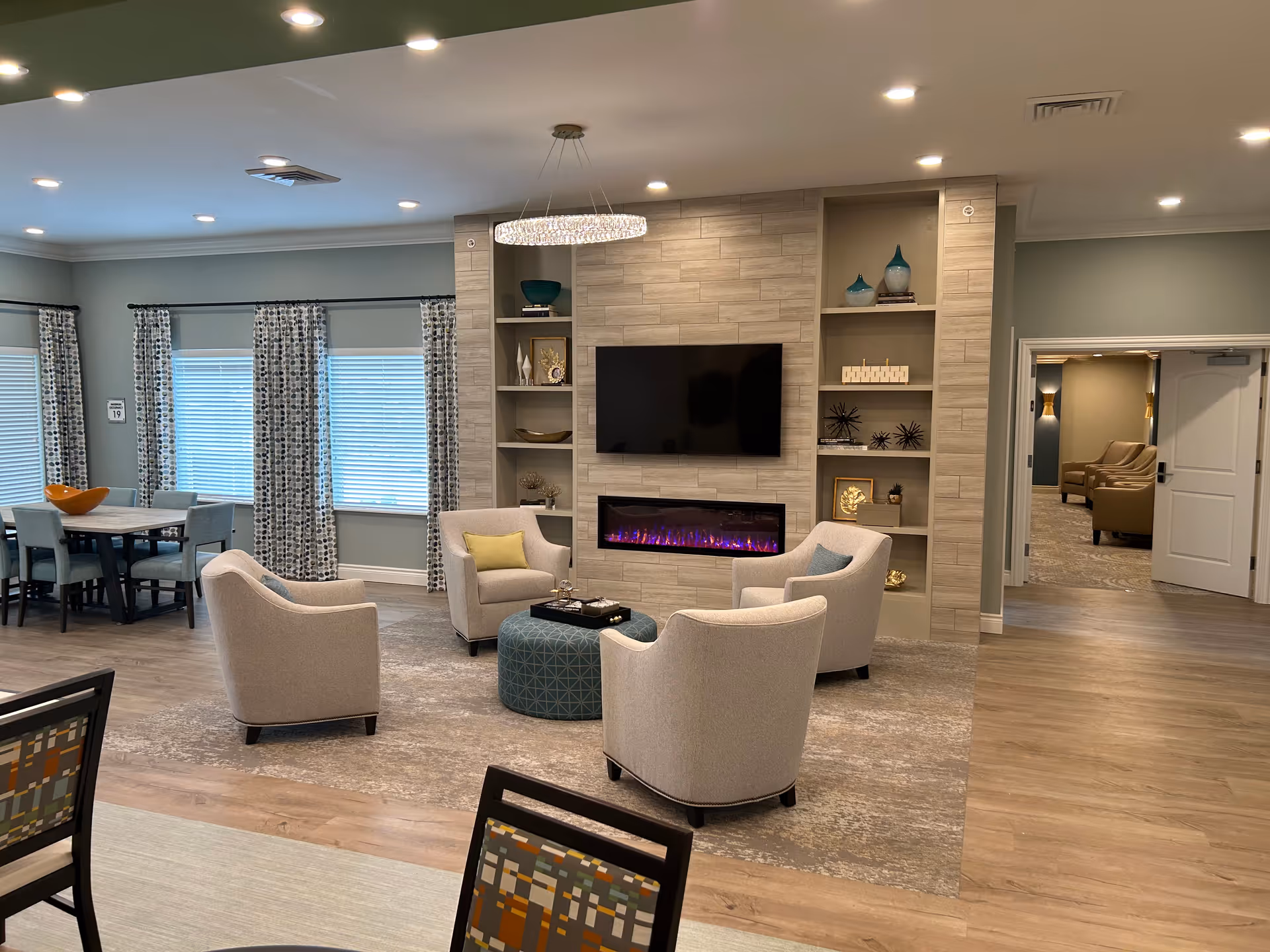 A modern and cozy living room area in a senior living facility featuring four beige armchairs arranged around a round blue ottoman. The room has a built-in wall unit with a flat-screen TV mounted above an electric fireplace with purple flames. Shelves on either side of the fireplace display decorative items. To the left, there is a dining area with a table and chairs near windows with patterned curtains. The room has wood flooring, recessed lighting, and a chandelier above the seating area. A doorway on the right leads to another room with additional seating.
