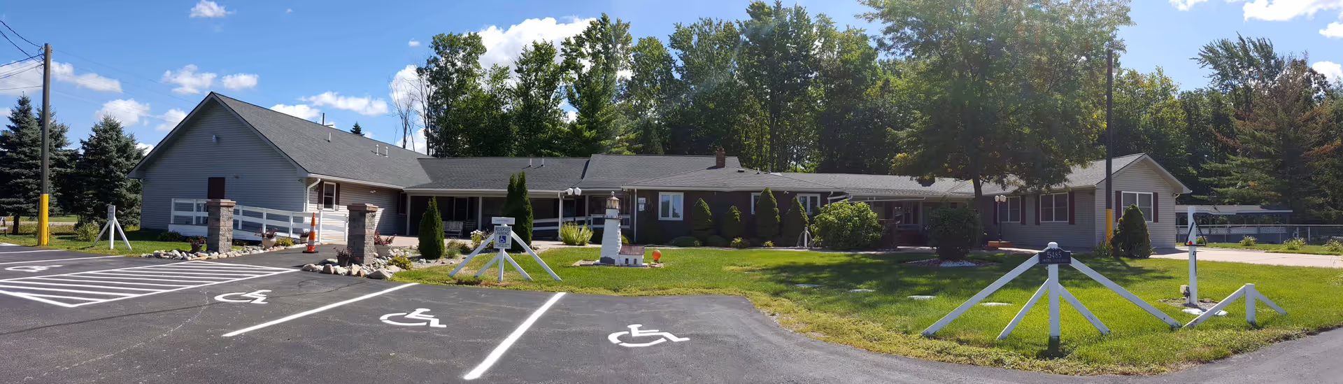 Single-story senior living facility building with a front lawn, handicap parking spaces, and trees in the background.