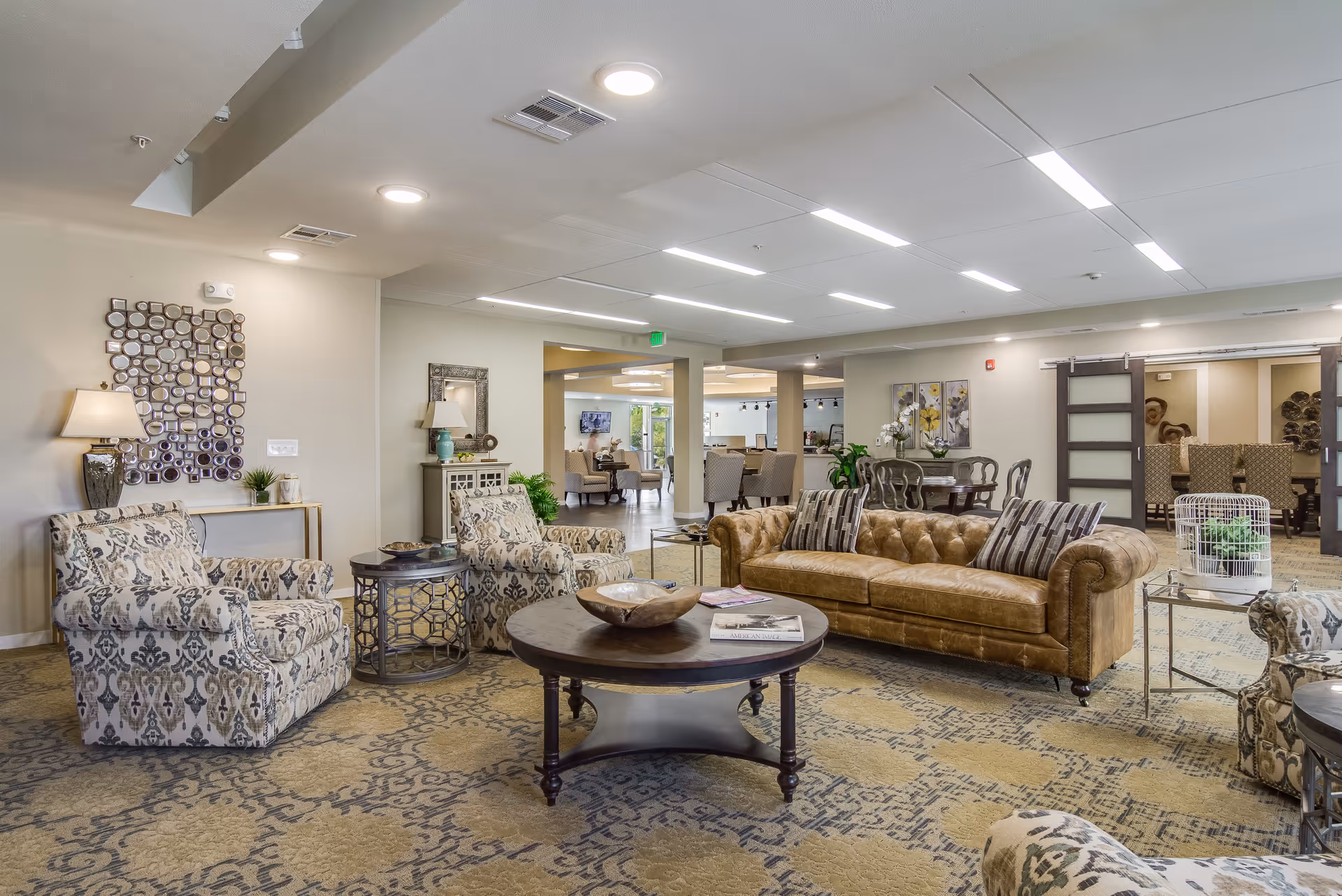 A spacious and well-lit living room area in a retirement facility featuring a brown leather sofa with striped cushions, two patterned armchairs, a round wooden coffee table with decorative items, and side tables with plants and lamps. The room has a patterned carpet, neutral-colored walls, modern ceiling lights, and an open layout leading to other seating and dining areas in the background.
