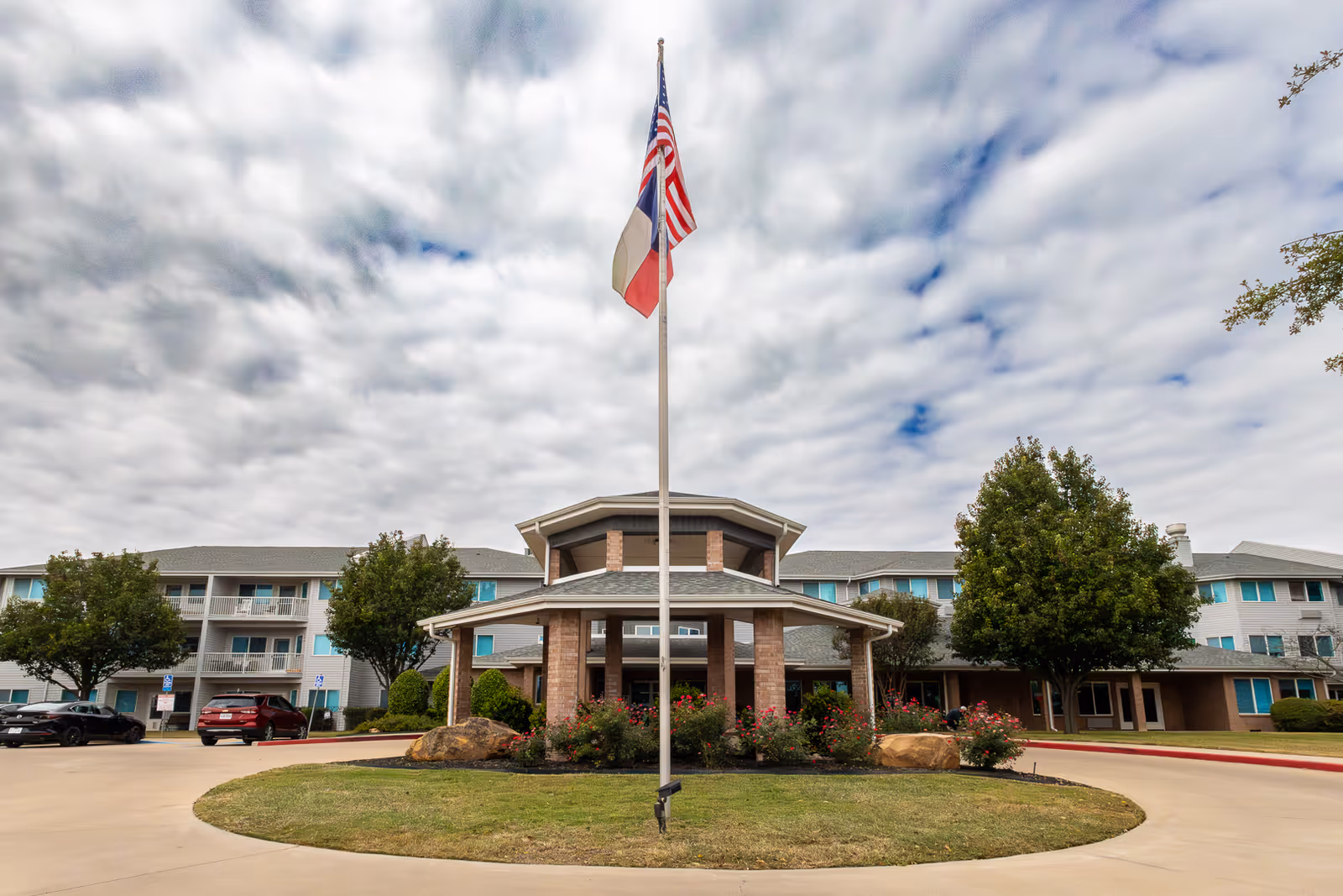 Front entrance of a multi-story senior living building with a flagpole flying U.S. and Texas flags and a circular driveway.