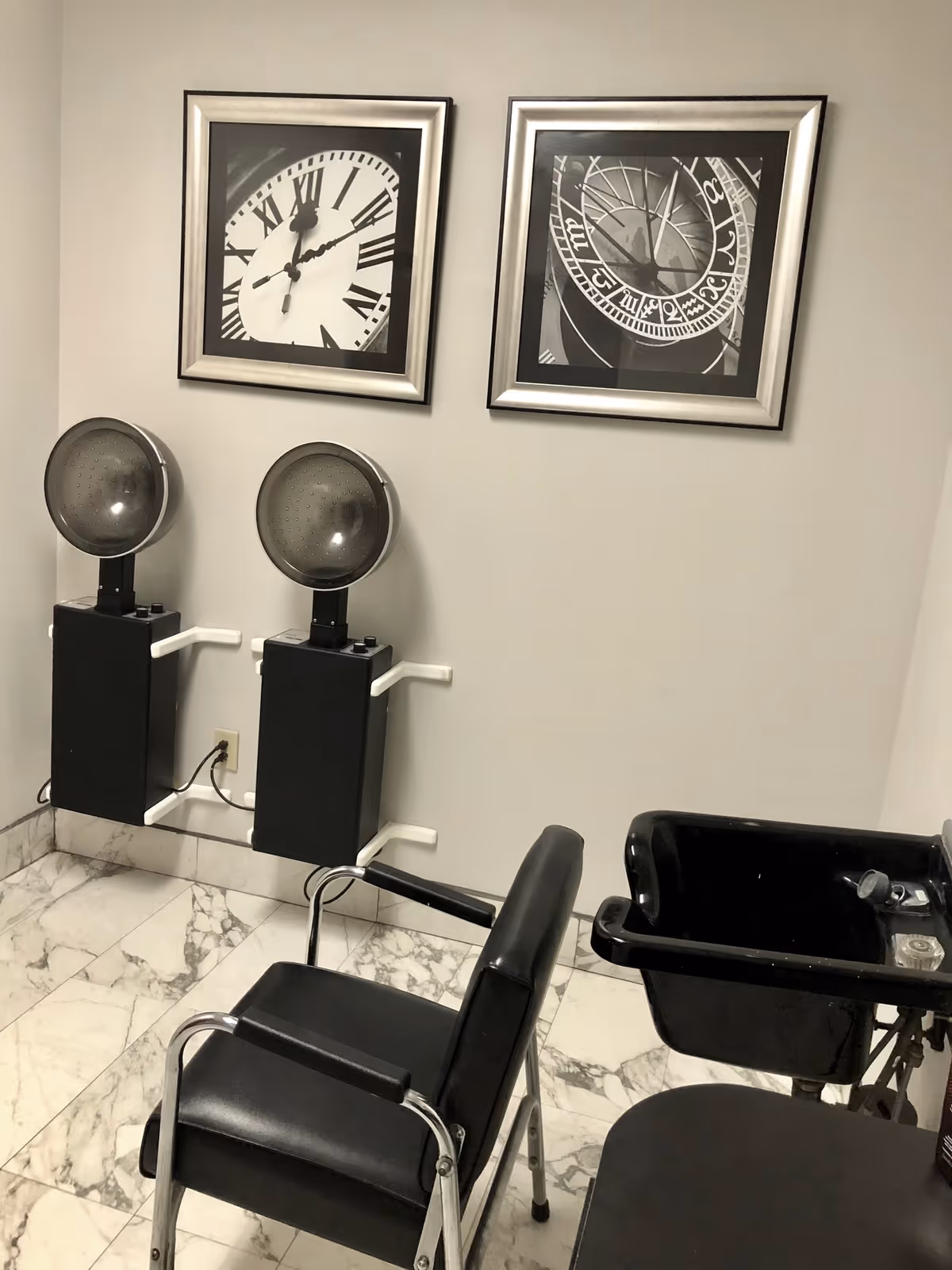 Interior salon area with two hooded hair dryers, a black salon chair and a wash basin beneath framed clock artwork on the wall.