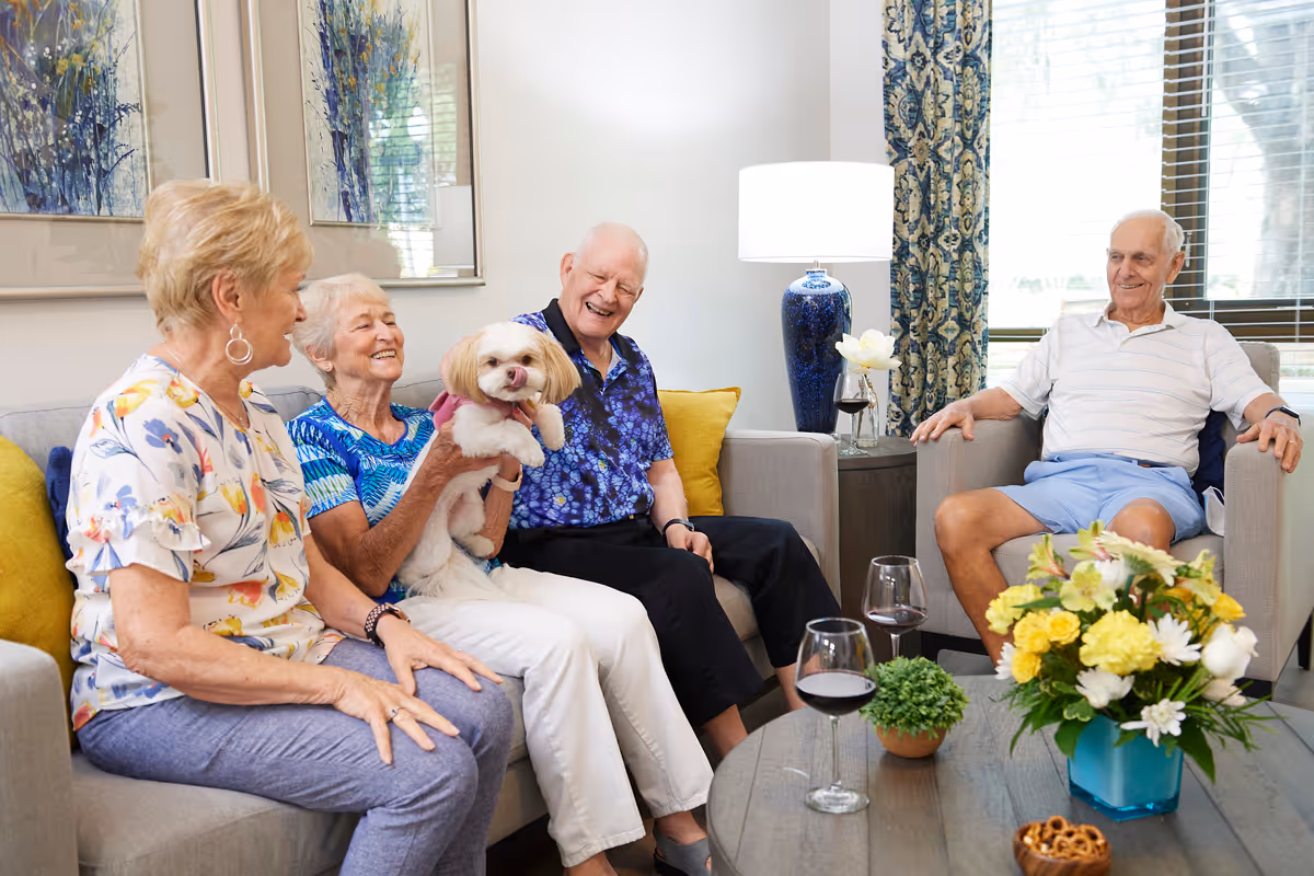 Four elderly people sitting in a living room, smiling and enjoying each other's company. One woman is holding a small white dog. There are two glasses of red wine on the coffee table, along with a small plant, a bowl of pretzels, and a vase of yellow and white flowers. The room has a cozy and bright atmosphere with framed artwork on the wall and a large window with patterned curtains.