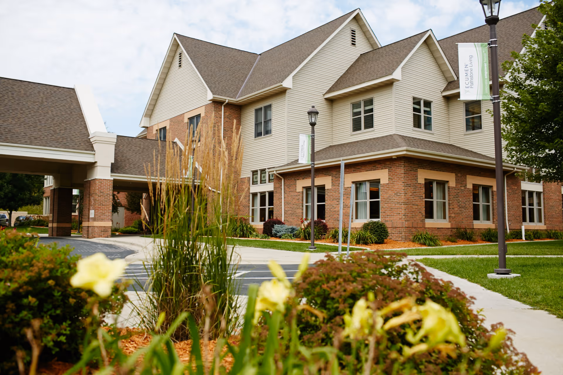 Exterior view of a two-story senior living facility building with brick and beige siding, surrounded by landscaped greenery and flowers. A covered entrance driveway is visible on the left, and lampposts with banners that read 'Ecumen Pathstone' are along the sidewalk.