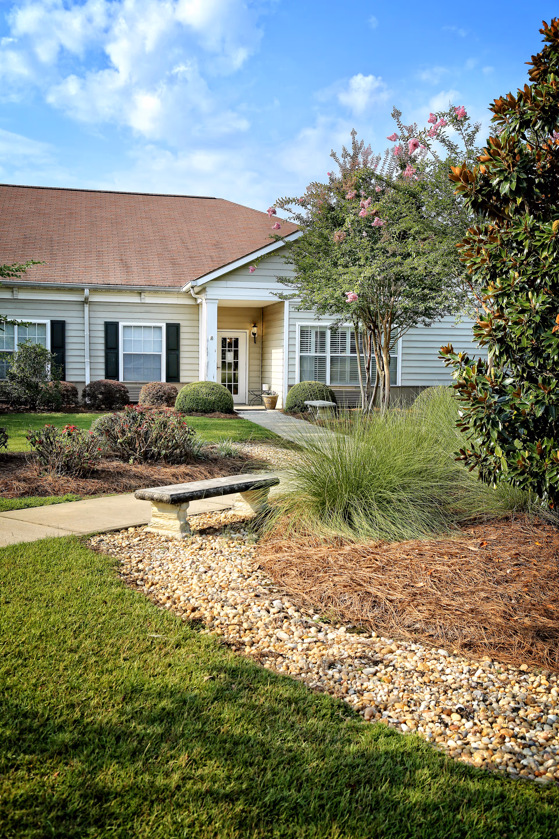 Front exterior of a single-story senior living building with a walkway, bench, and landscaped lawn and shrubs.