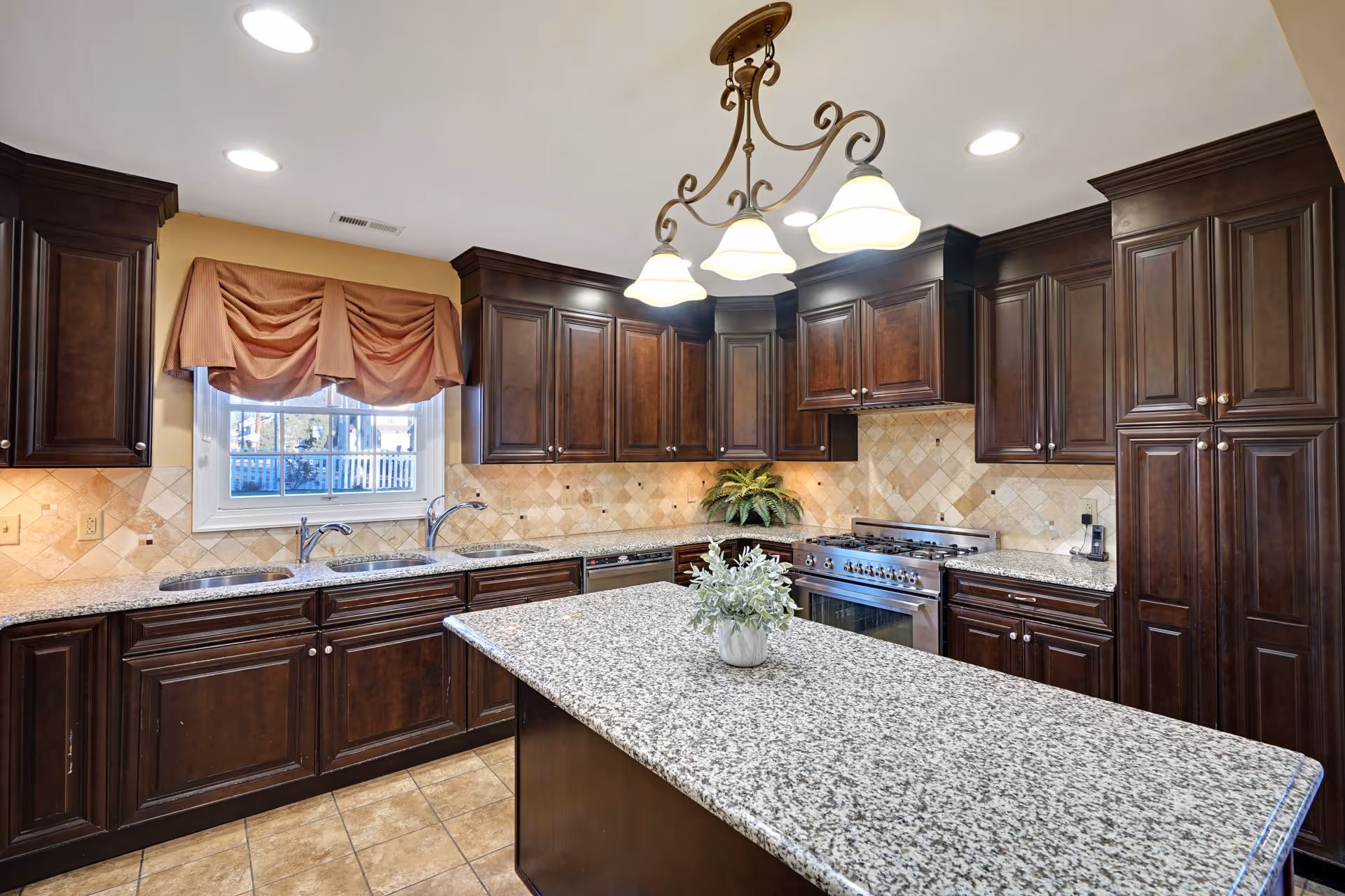 A spacious kitchen with dark wooden cabinets, granite countertops, and a central island with a small potted plant. The kitchen features a double sink under a window with a red valance, a stainless steel stove with an oven, and a decorative tile backsplash. A chandelier with four lights hangs from the ceiling.