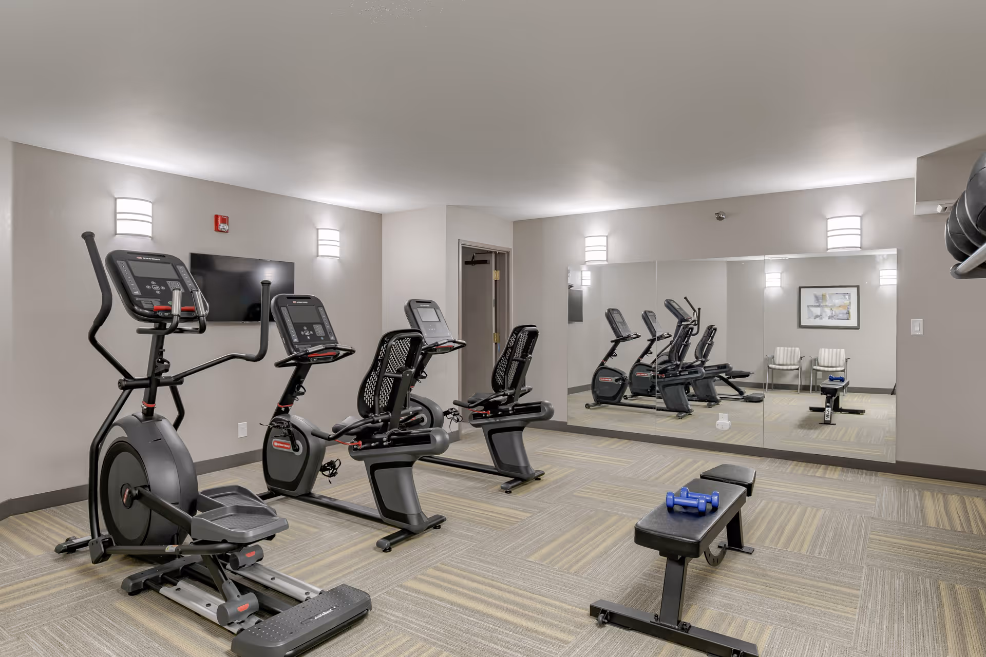 A small fitness room with exercise equipment including two recumbent bikes, an elliptical machine, a weight bench with blue dumbbells, a large wall mirror, and a wall-mounted TV. The room has beige walls, carpeted flooring, and modern wall lights.