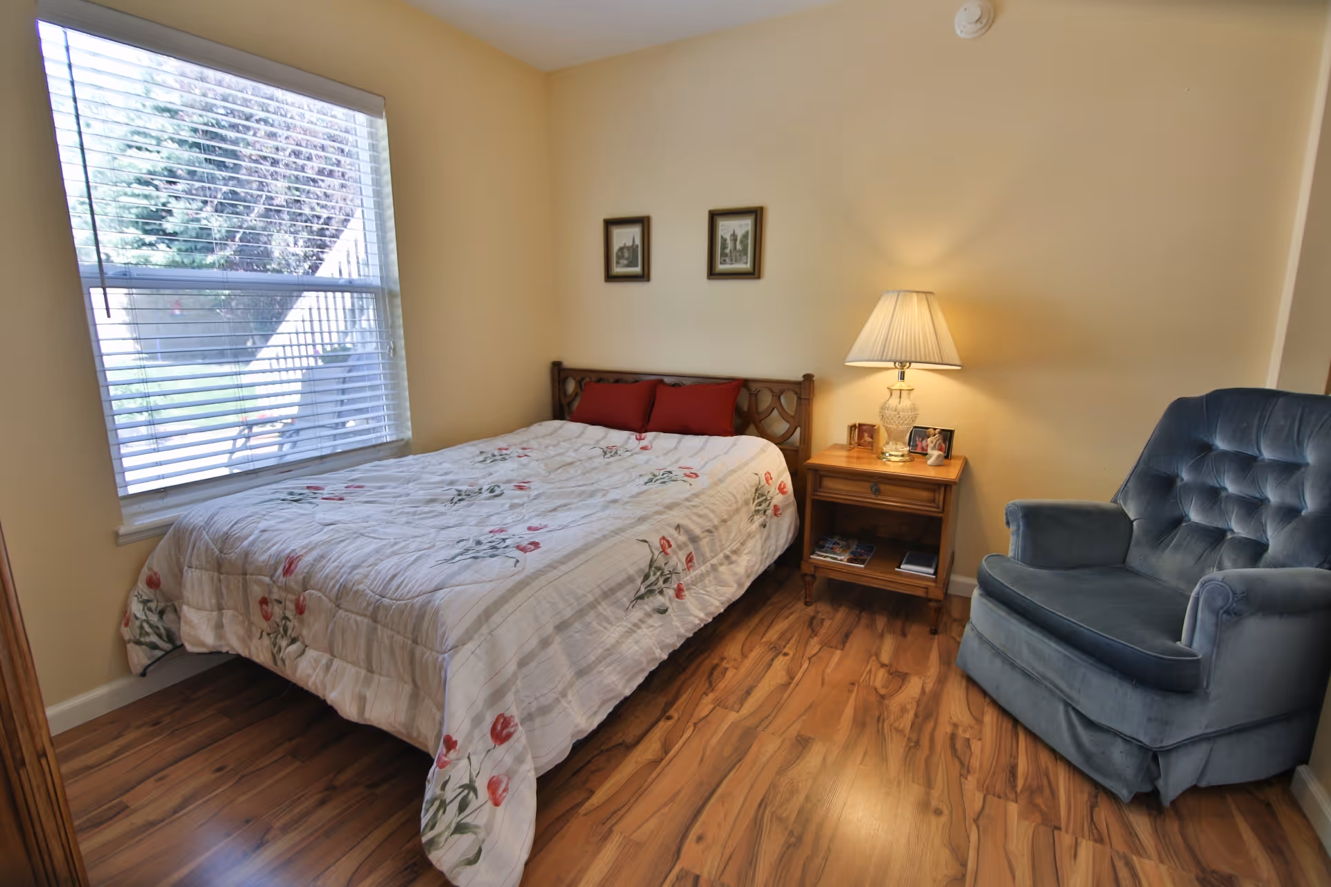 A cozy bedroom with a bed covered in a floral quilt, two red pillows, a wooden nightstand with a lamp and framed photos, a blue upholstered armchair, and a window with white blinds letting in natural light.