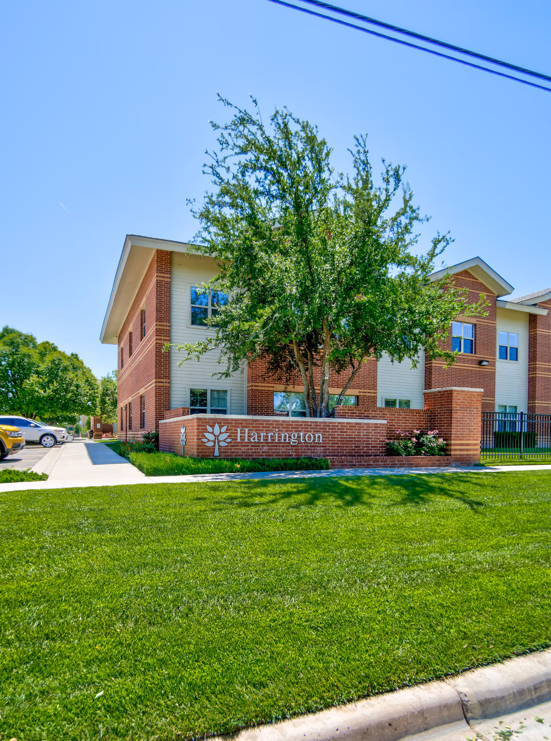 Exterior view of a two-story brick and siding building with a green lawn and a tree in front. A brick sign in front of the building reads 'Harrington'. Several parked cars are visible on the left side under a clear blue sky.
