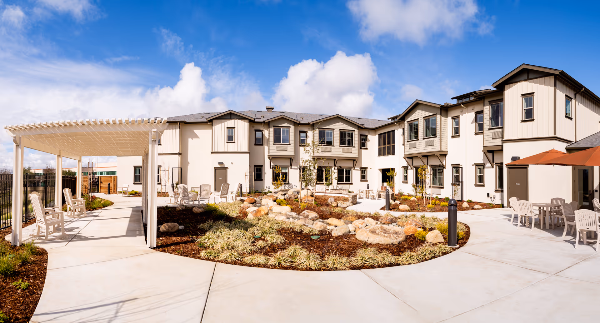 Outdoor courtyard area of a senior living facility with a two-story building in the background. The courtyard features a paved walkway, seating areas with chairs and tables under umbrellas, a pergola with chairs underneath, landscaped garden beds with rocks and plants, and a bright blue sky with some clouds.