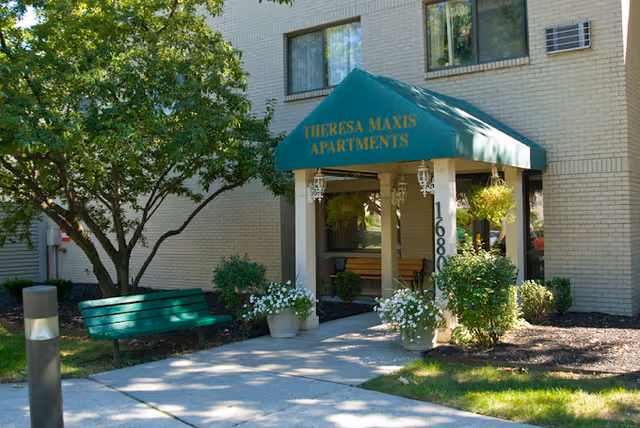 Entrance to Theresa Maxis Apartments with a green awning displaying the name. The building is light-colored brick with windows, surrounded by greenery including trees, bushes, and potted plants. A green bench is visible on the left side of the walkway leading to the entrance.