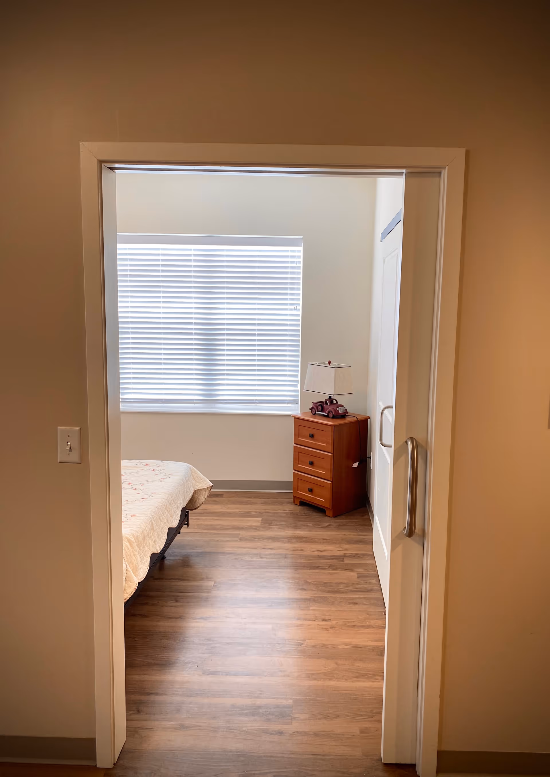 View through a doorway into a bedroom with wooden flooring, a bed partially visible on the left, a wooden nightstand with three drawers on the right, and a lamp with a red toy car base on top. A window with closed white blinds is centered on the back wall.