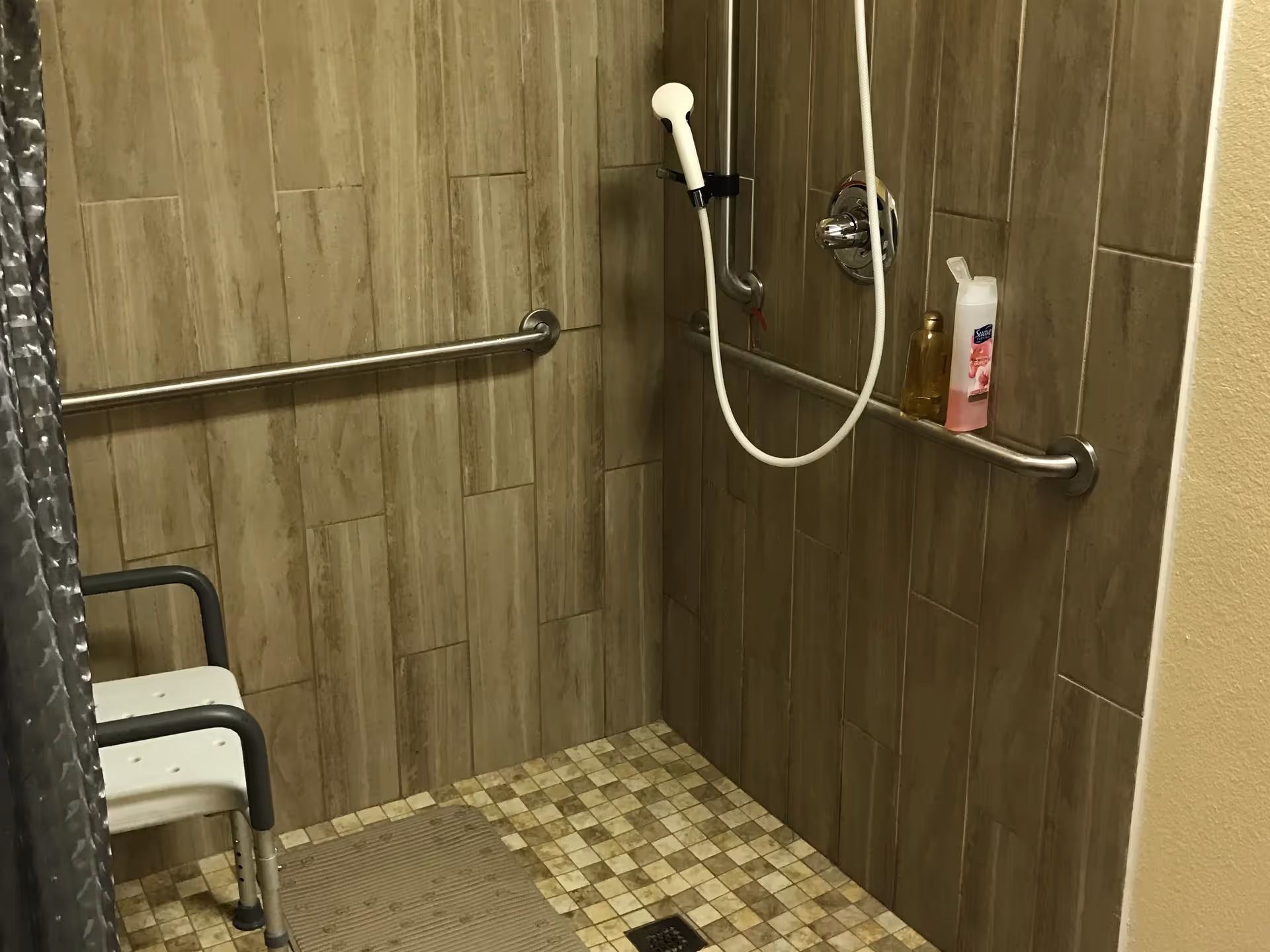 Accessible tiled shower with grab bars, a handheld shower head, a shower chair, and toiletries on a wall rail.
