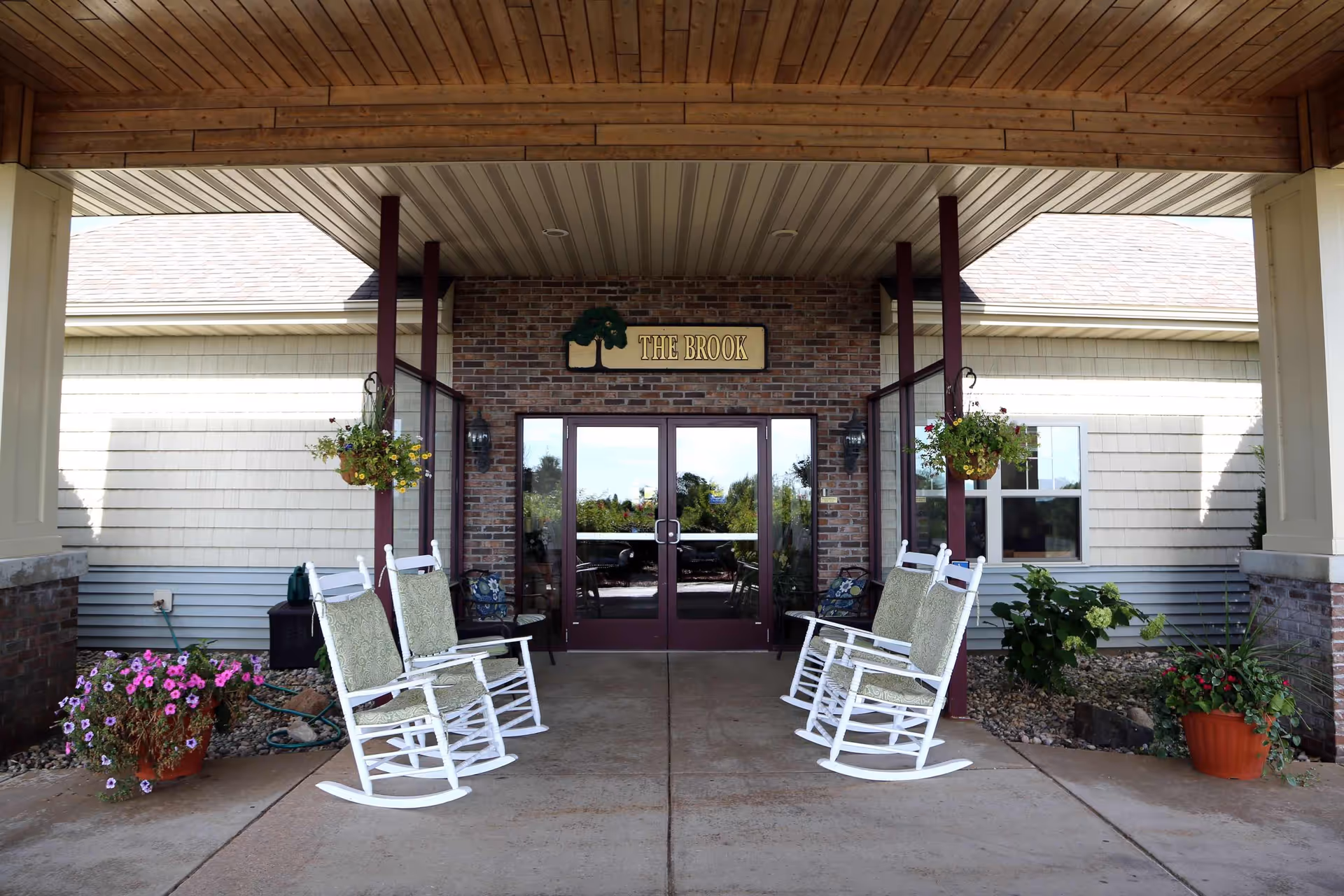 Entrance of The Brook senior living facility with a covered porch featuring four white rocking chairs with cushions, hanging flower baskets, potted plants, and double glass doors under a sign that reads 'THE BROOK'.