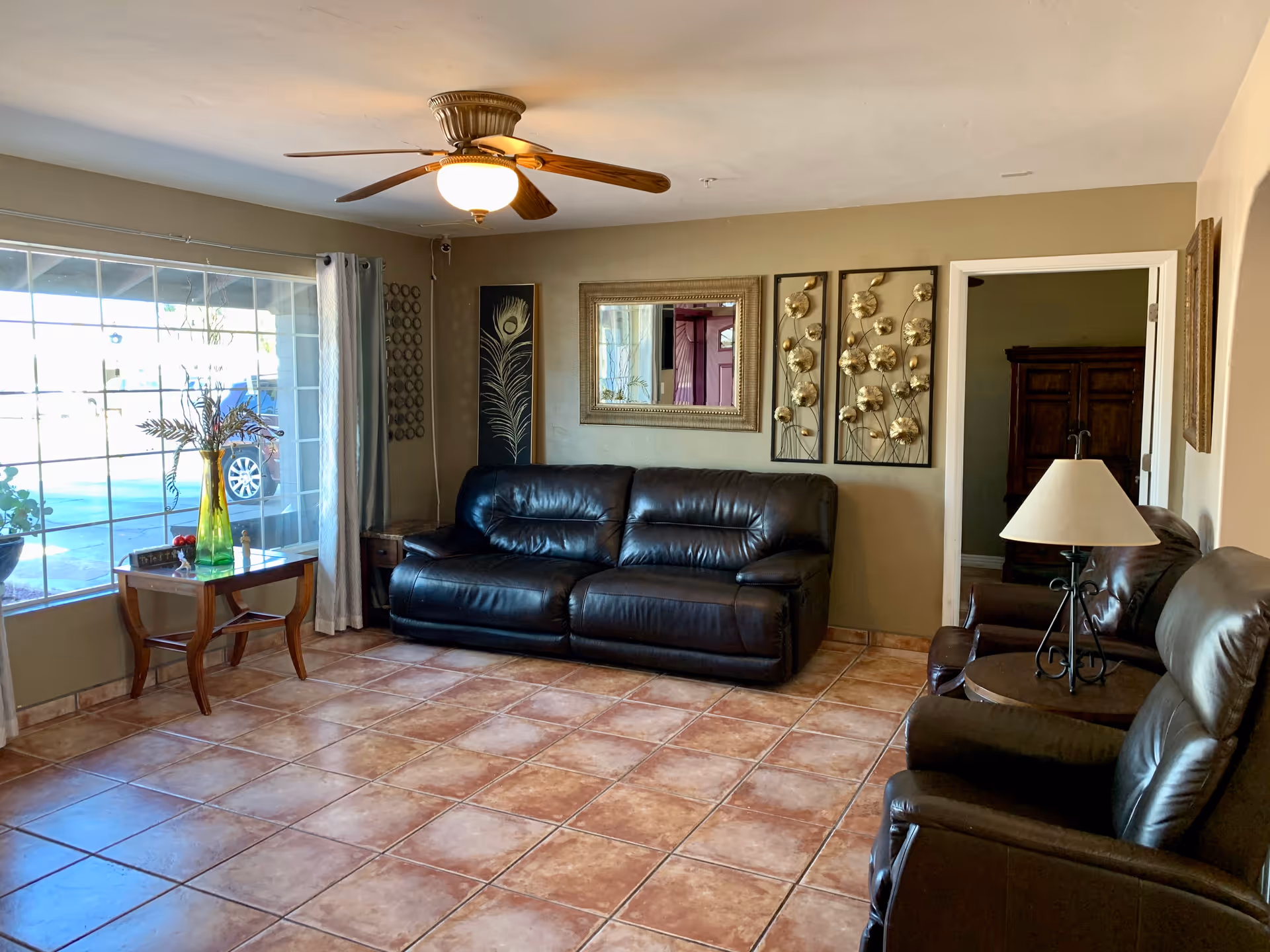 A living room with a large window on the left side letting in natural light. The room features a black leather sofa and two matching armchairs. There is a wooden side table with a green vase holding decorative branches near the window. The walls are decorated with framed artwork and a mirror. A ceiling fan with a light fixture is mounted on the ceiling. The floor is tiled with brown and beige tiles.