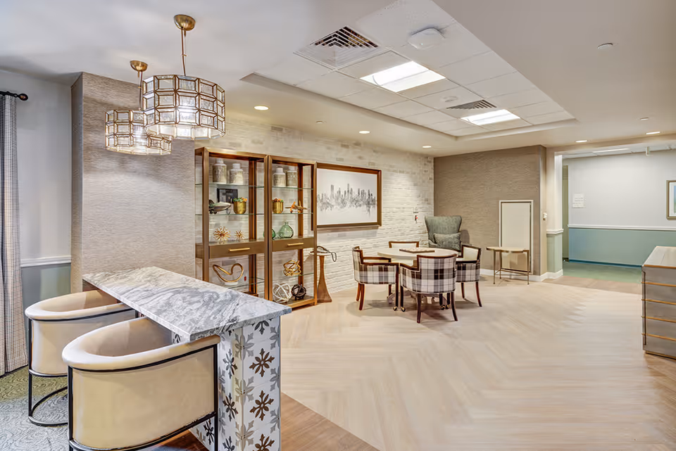 Bright senior living common area with a marble-topped bar, pendant lights, a seating area with plaid chairs around a table, and decorative shelving.