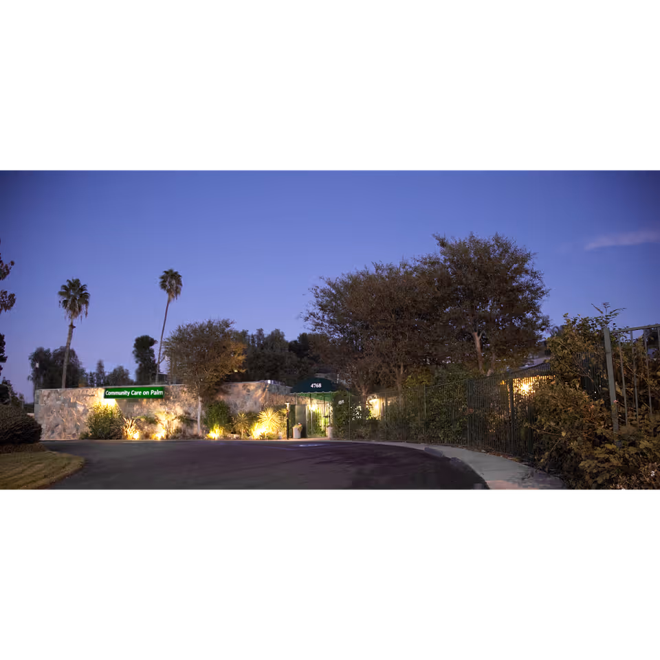 Exterior view of Community Care On Palm nursing facility in Riverside during dusk, showing a stone wall with a green sign, palm trees, other trees, and a driveway leading to the entrance.