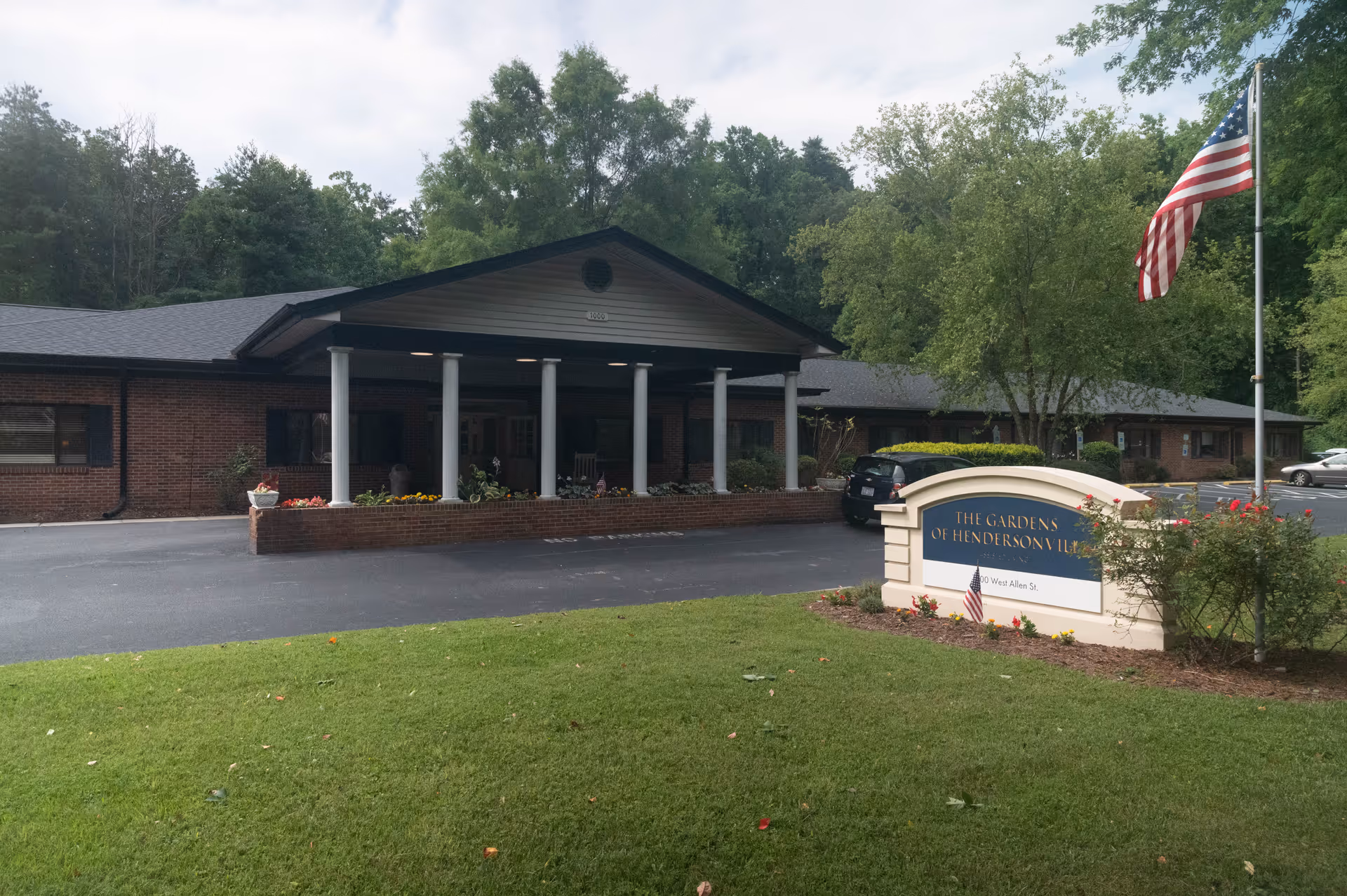 Exterior view of The Gardens of Hendersonville facility showing a single-story brick building with a covered entrance supported by white columns, a parking area with cars, a green lawn, an American flag on a flagpole, and a sign with the facility's name.
