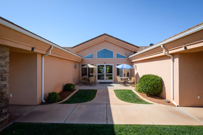 Outdoor courtyard area of a senior living facility with beige walls, a concrete pathway, green bushes, and patio tables with umbrellas near the entrance door under a clear blue sky.