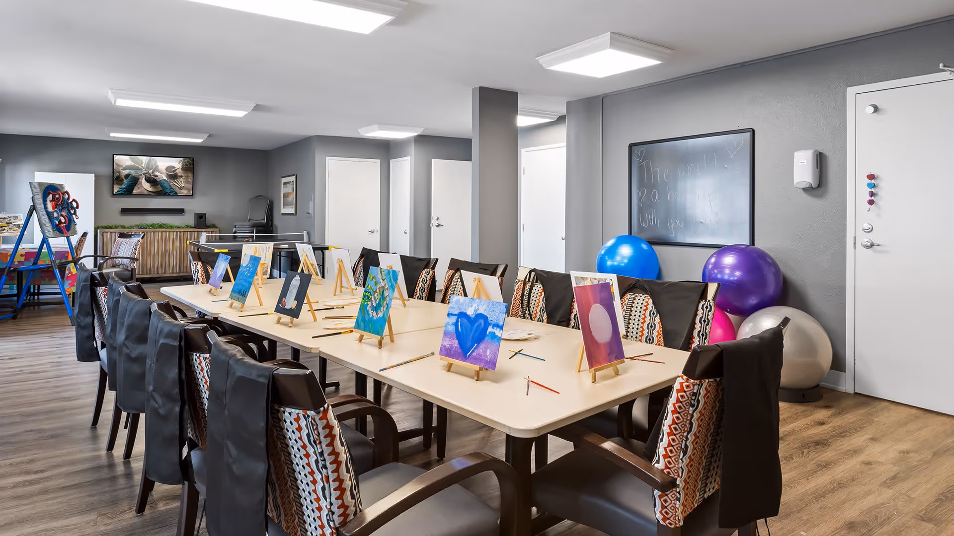 A bright activity room with a long table surrounded by chairs, each chair having a small canvas on an easel with colorful paintings and paintbrushes. The room has wooden flooring, gray walls, a blackboard, exercise balls in the corner, and a TV mounted on the wall. There are also easels and art supplies visible in the background.