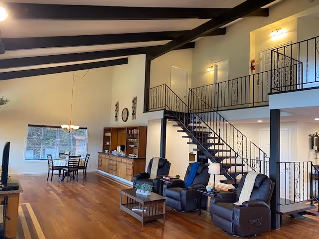 Spacious interior of a senior living facility with wooden floors, a dining area with a table and chairs near a window, a wooden cabinet along the wall, and a seating area with three recliner chairs and a coffee table. A staircase with black metal railings leads to an upper level with doors and wall-mounted lights.