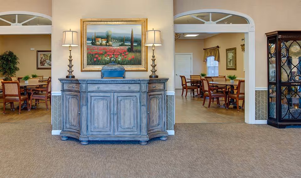 Interior view of a senior living facility showing a hallway with a wooden sideboard topped with two lamps and a framed painting of a countryside scene with red flowers. On either side of the hallway, there are open archways leading to dining areas with tables, chairs, and plants. The walls are beige with white trim and the floor is carpeted in the hallway and wood in the dining areas.