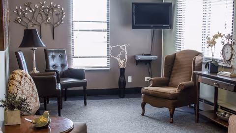 A cozy sitting room with armchairs, a wall-mounted TV, side tables, and decorative accents near windows.
