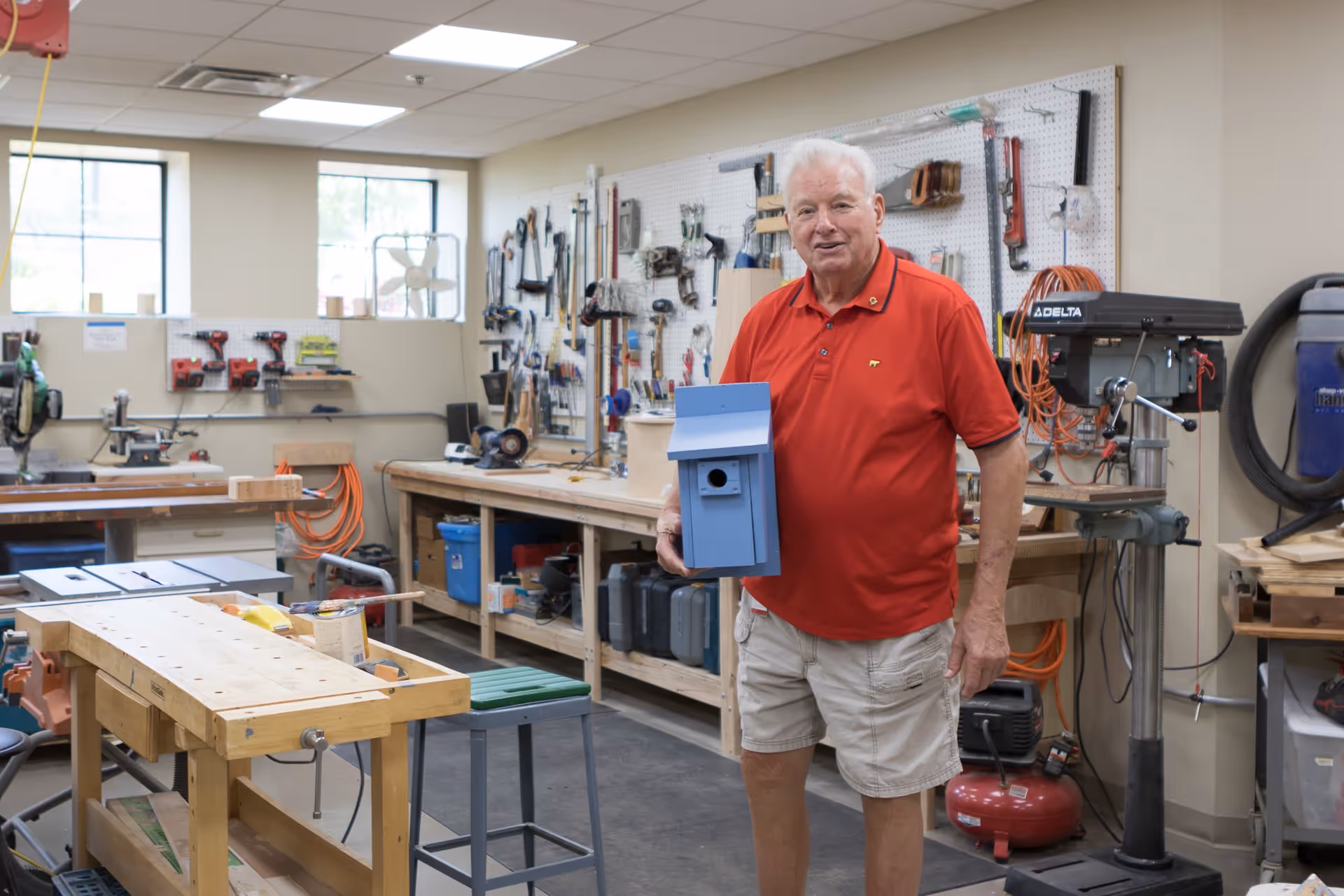 An elderly man wearing a red polo shirt and beige shorts stands in a woodworking workshop holding a blue birdhouse. The workshop has various tools hanging on a pegboard, workbenches, and equipment such as a drill press and saws. There are windows letting in natural light.