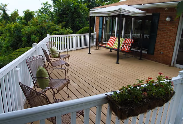 Outdoor wooden deck with white railing, featuring three metal chairs with green cushions and a canopy swing with colorful striped pillows. There is a flower box with blooming flowers attached to the railing, and trees and greenery are visible in the background.