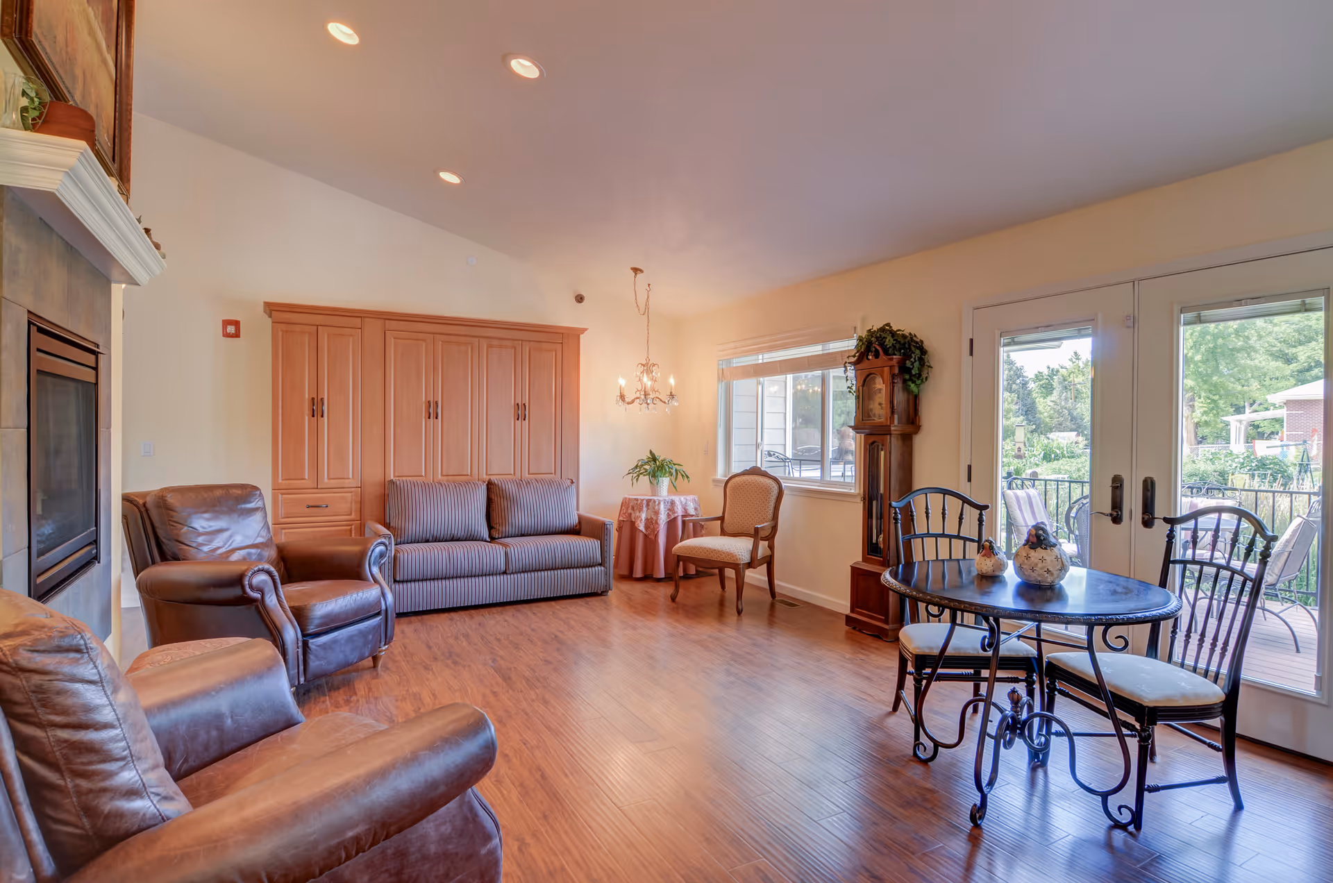 A bright and spacious living room with wooden flooring, featuring two brown leather armchairs, a striped sofa, a small round table with a pink tablecloth and a plant, a wooden grandfather clock, and a round black metal table with three chairs near glass doors leading to an outdoor patio.