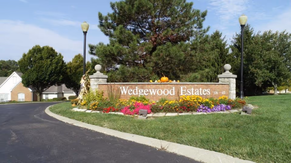 Entrance sign for Wedgewood Estates surrounded by colorful flowers and greenery, with a paved road curving to the left and trees in the background under a partly cloudy sky.