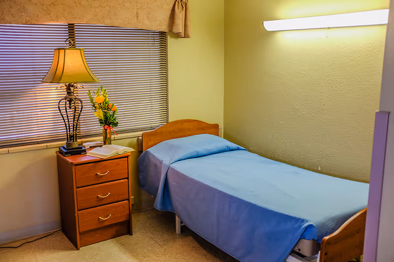 A small bedroom with a single bed covered in a blue blanket, a wooden nightstand with three drawers, a table lamp, a vase with flowers, and a window with closed blinds and a valance.