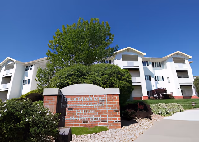 Exterior view of a senior living facility with white multi-story buildings, green bushes, trees, and a clear blue sky. A brick sign structure is in the foreground, partially obscured by greenery.