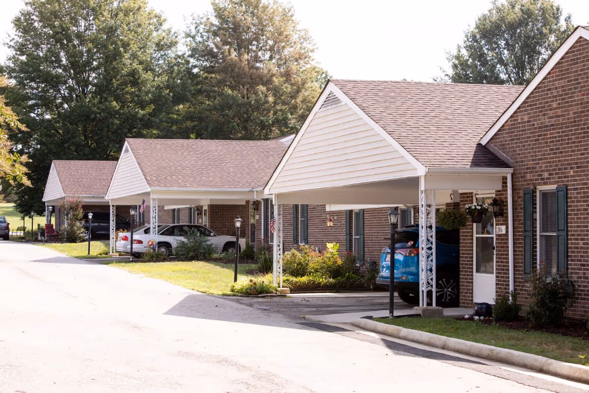 A row of single-story brick residential units with covered carports, each with a pitched roof and white trim. Several cars are parked under the carports, and there are small landscaped areas with bushes and flowers in front of the units. Trees and a grassy area are visible in the background.