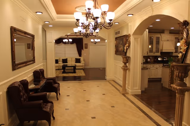 Interior view of a senior living facility showing a hallway with beige tiled floor and two brown leather chairs on the left side. The hallway leads to a living area with a white sofa and a dark coffee table. On the right side, there is an arched entrance to a kitchen with white cabinetry and dark countertops. The ceiling features recessed lighting and two ornate chandeliers. Decorative columns and framed artwork adorn the walls.