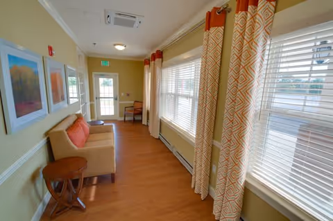 A bright hallway with large windows on the right side covered with white blinds and orange patterned curtains. On the left side, there is a beige loveseat with orange cushions, a small round wooden side table, and three framed artworks on the wall. At the end of the hallway, there is a door with a window and an exit sign above it. The floor is wooden, and the walls are painted light yellow.