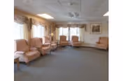 A spacious living room area with multiple beige upholstered armchairs arranged along the walls. The room has large windows with valance curtains allowing natural light to enter. There is a framed picture on the wall and a side table with a lamp near one of the chairs. The floor is carpeted in a neutral color.